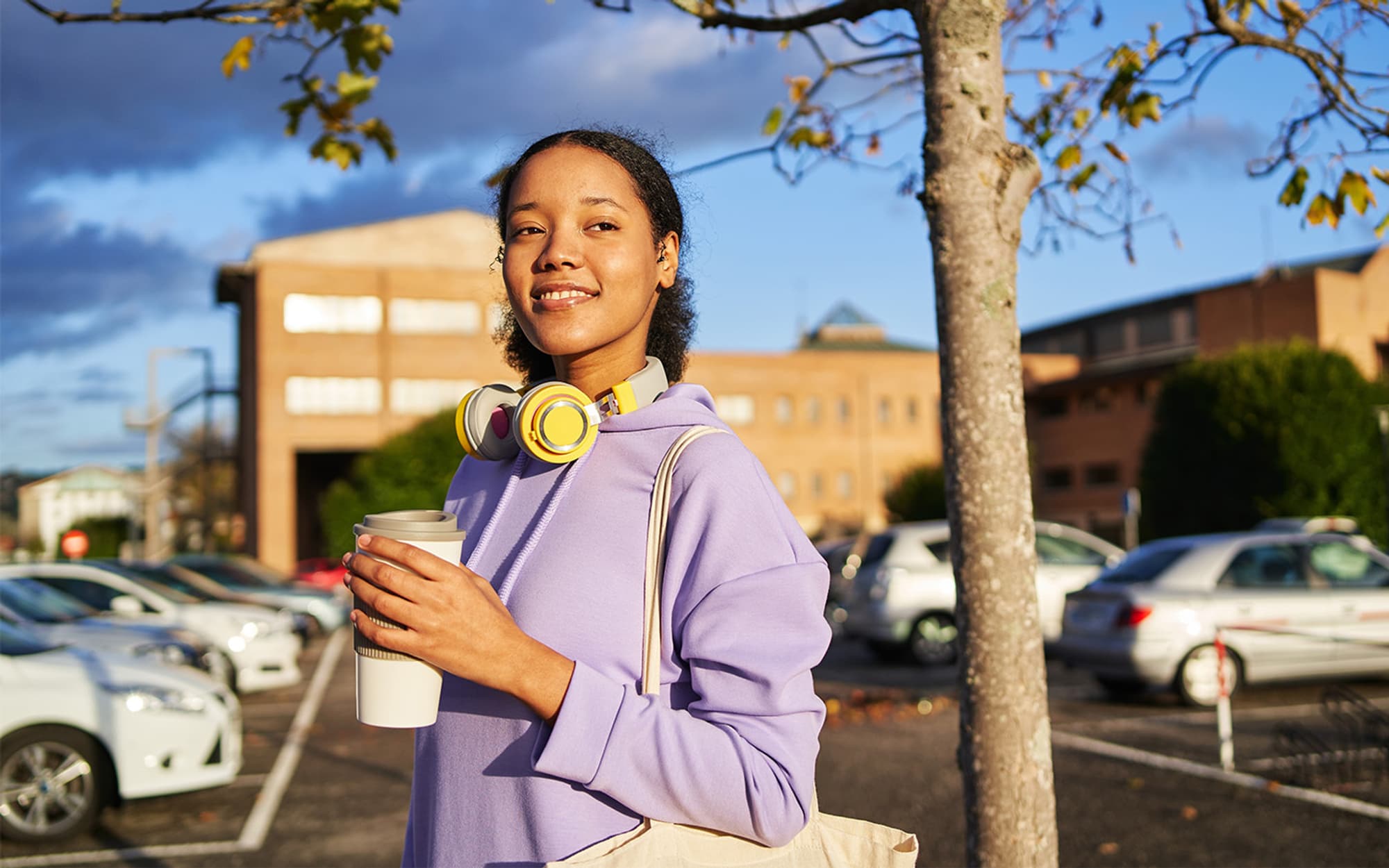A young mixed-race woman in purple workout gear standing in a carpark holding a grey reusable coffee cup.