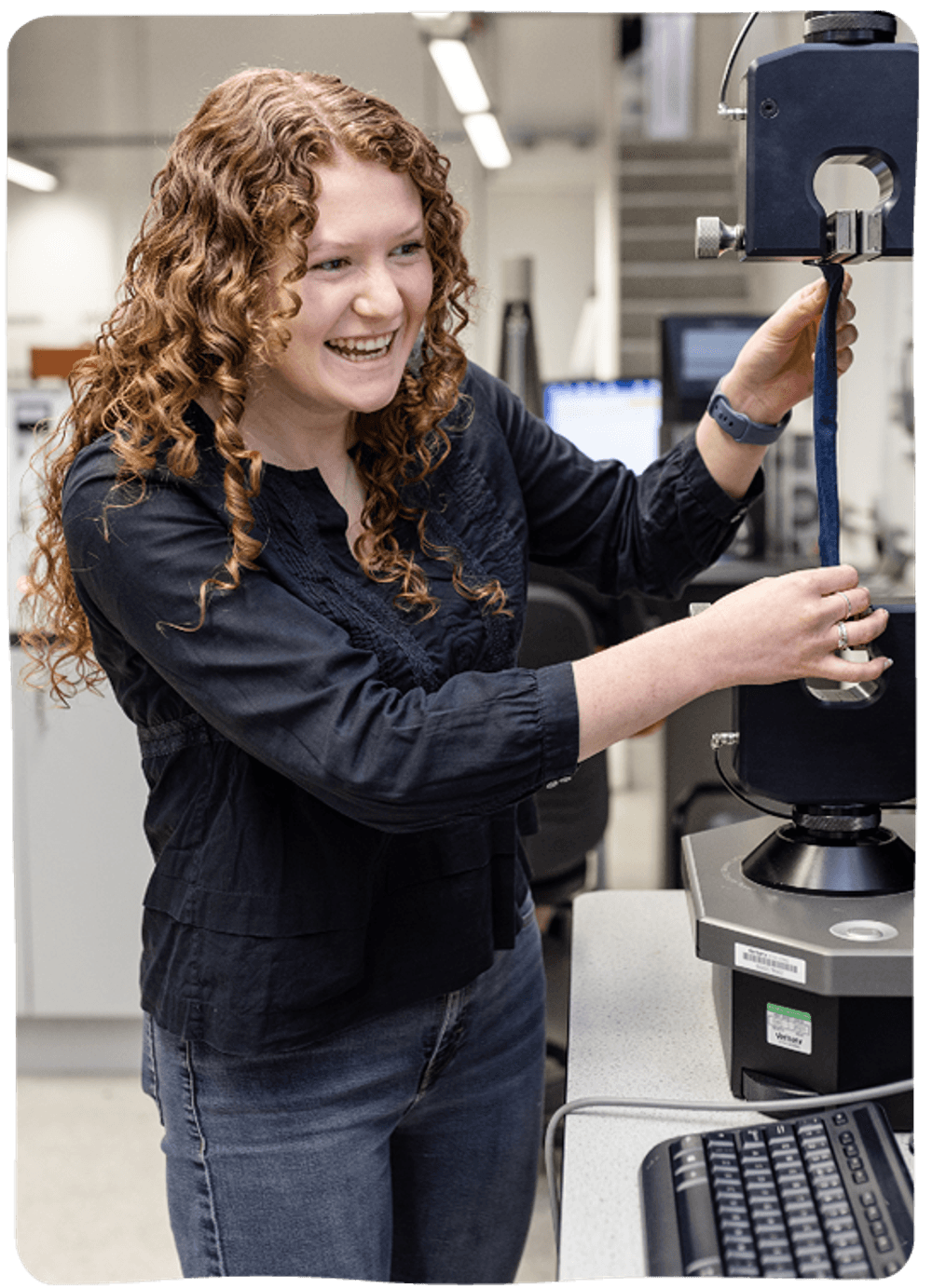 A young woman with curly hair is standing smiling while operating a machine in a lab.
