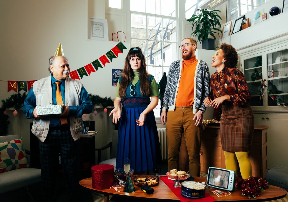 Four office workers wearing colourful 80s clothing stand in an office decorated for Christmas. From left to right: an older gentleman in a shirt and tie, the office boss, holds a gift and looks at the other three; a woman in her 30s shrugs at the camera and rolls her eyes; a man in his late 30s looks at the boss in a confused way, while laughing; a young woman laughs while looking at the other three.
