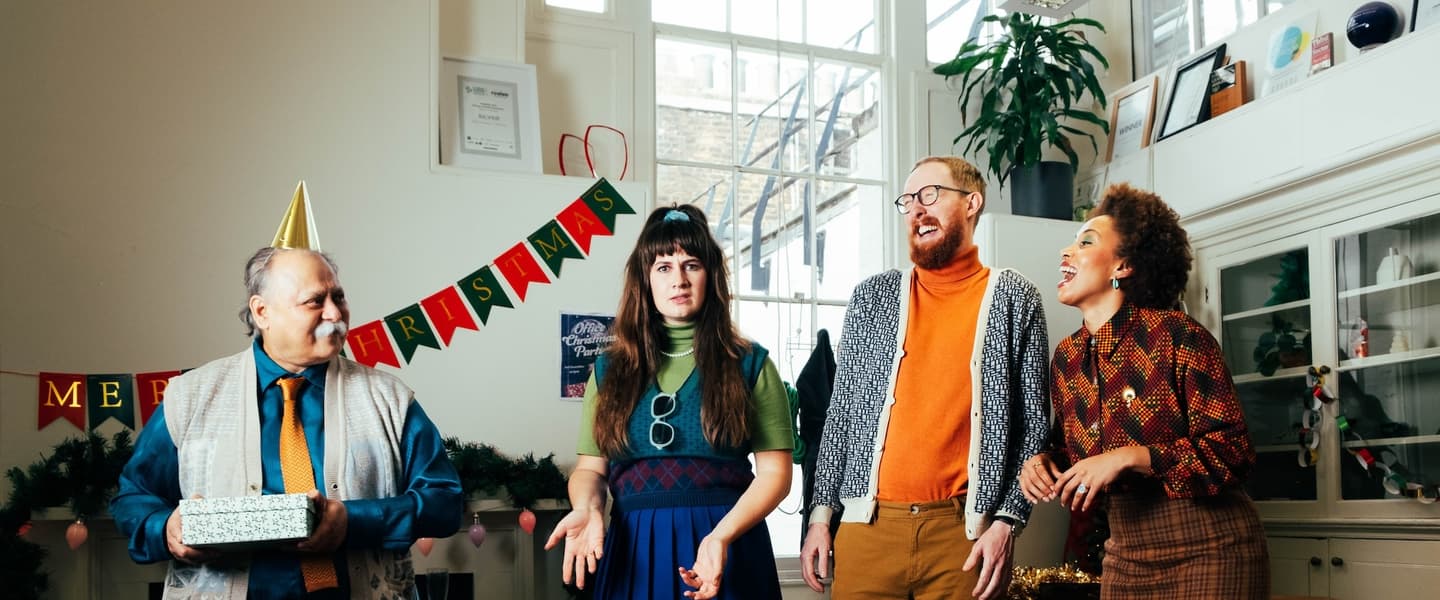 Four office workers wearing colourful 80s clothing stand in an office decorated for Christmas. From left to right: an older gentleman in a shirt and tie, the office boss, holds a gift and looks at the other three; a woman in her 30s shrugs at the camera and rolls her eyes; a man in his late 30s looks at the boss in a confused way, while laughing; a young woman laughs while looking at the other three.