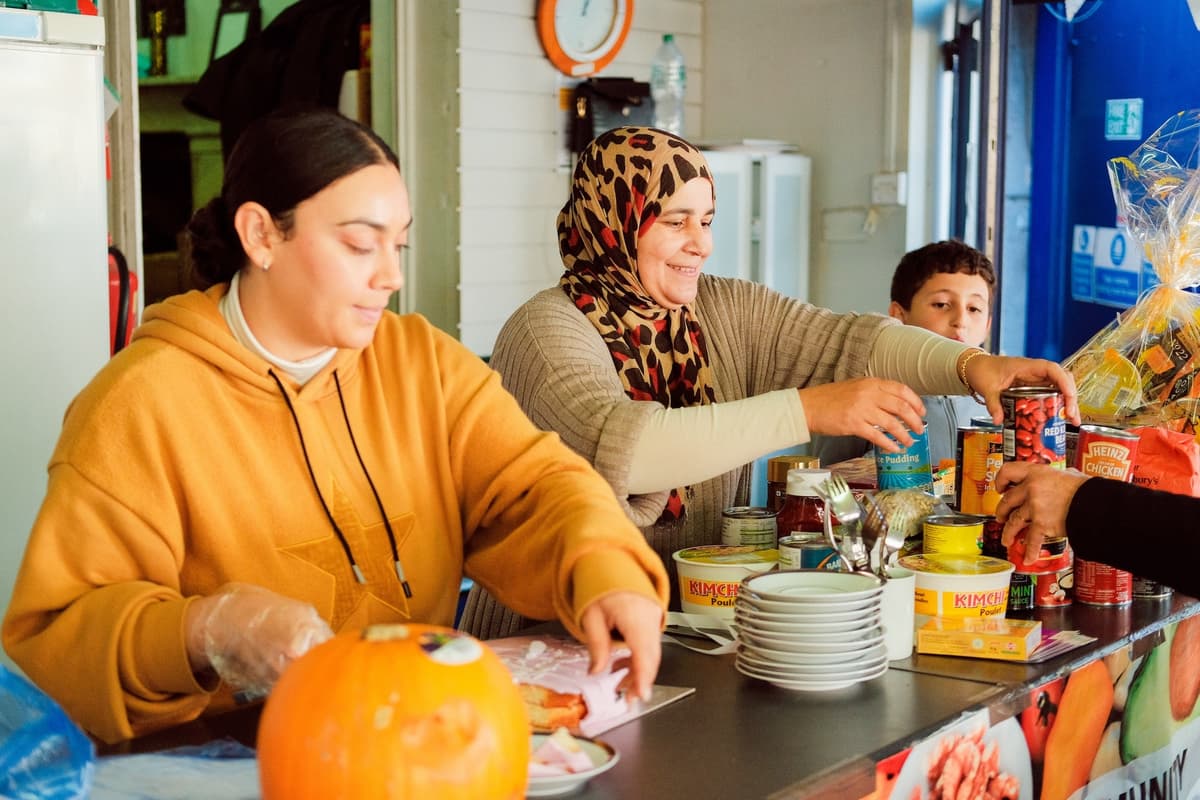 A young woman and an older women wearing a leopard-print hijab stand side by side behind a kitchen bench, and a young boy watched on besides them. They are handling food and a pile of plates is on the counter.