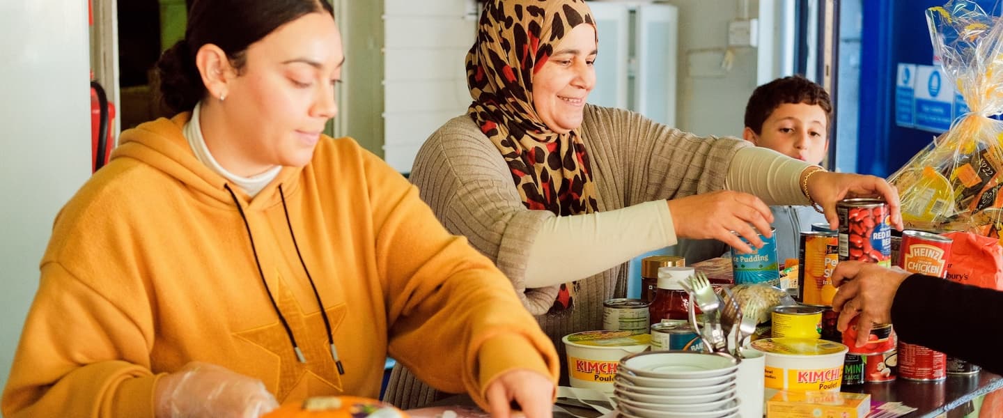 A young woman and an older women wearing a leopard-print hijab stand side by side behind a kitchen bench, and a young boy watched on besides them. They are handling food and a pile of plates is on the counter.