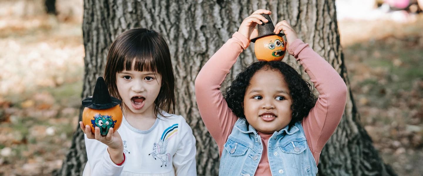Two young girls show off painted pumpkins. The small pumpkins they're holding each have a spooky face painted on, and don a witch hat.