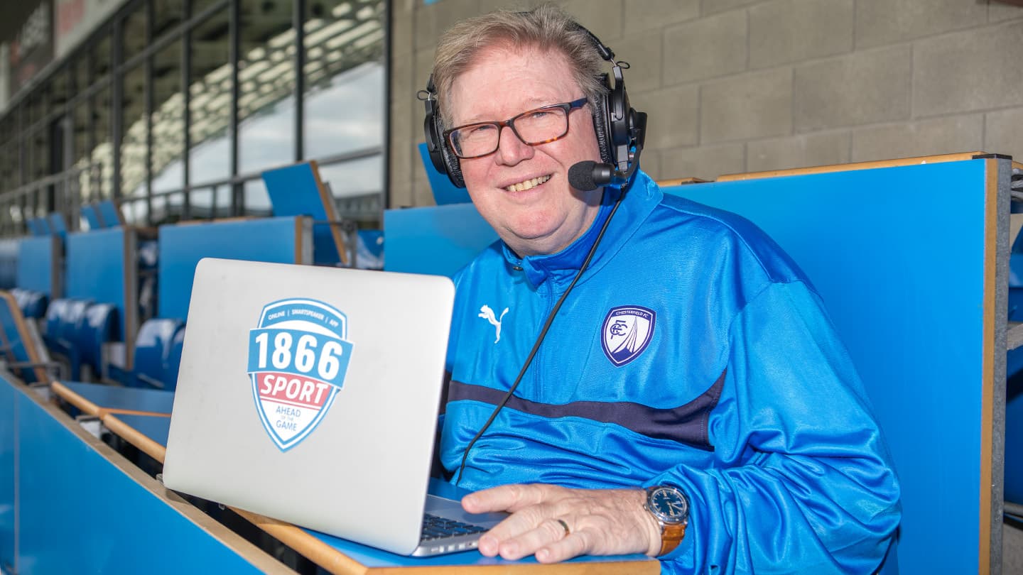A sports presenter looking at the camera, with a laptop and a headset, sitting in the football ground of Chesterfield FC.