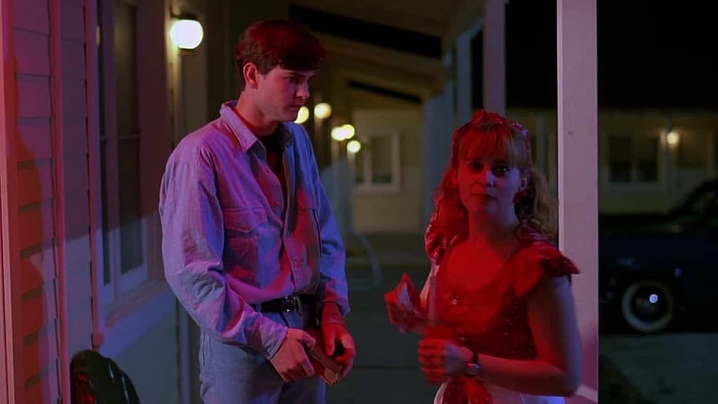 The lighting is red and purple. On the left, a male white teenager with brown hair looks towards a female white teenager with blonde hair and blue eyes. She looks at the camera.