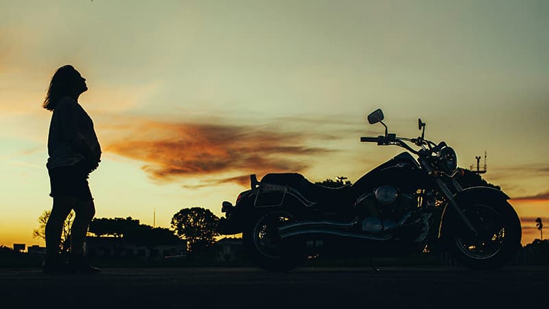A silhouetted woman and her motorbike, against a sunset background.