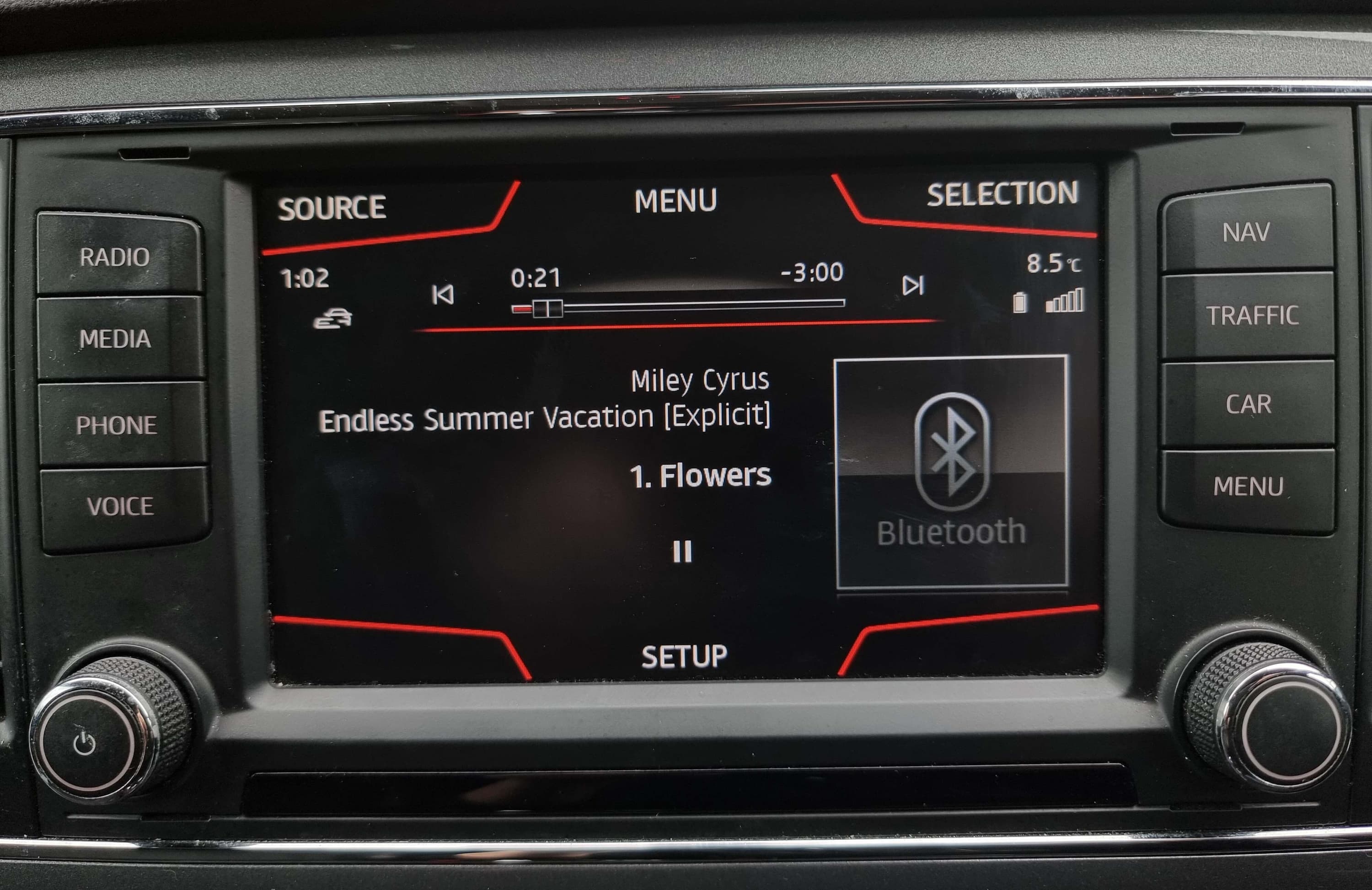 The screen and buttons of an car stereo. The screen shows a song playing and the Bluetooth symbol shows tis on the right hand side of the screen.