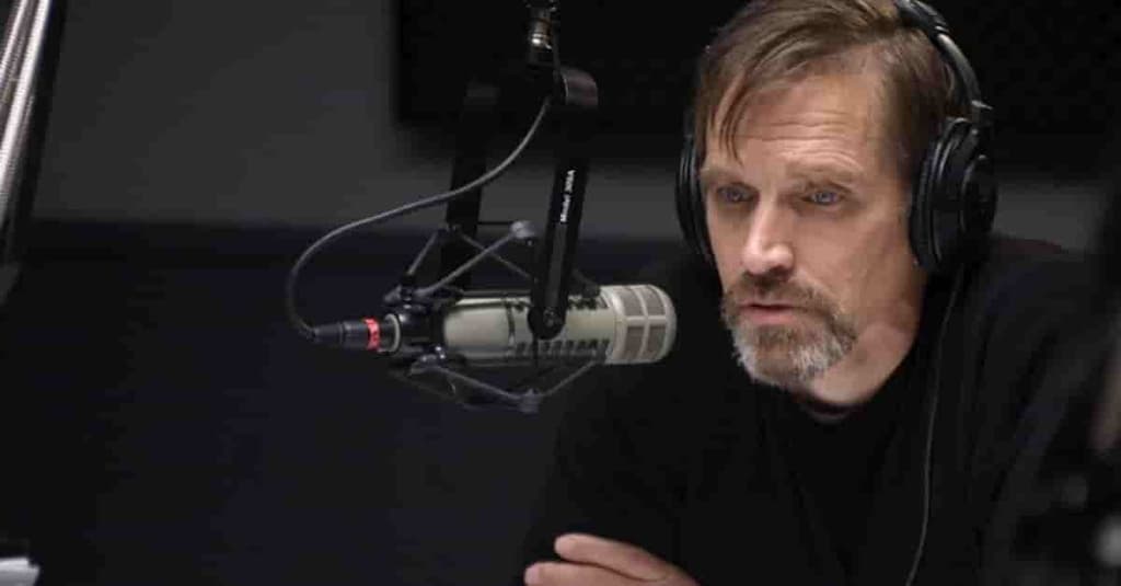 A white man in his mid 50s presents radio. He is looking solemn as he leans in front of the microphone. He has a peppered beard and grey hair.