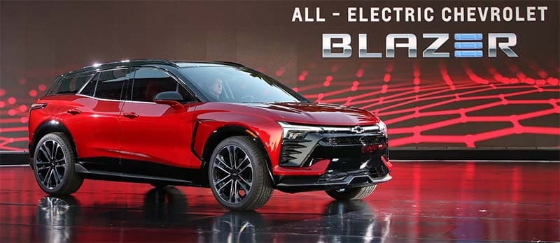 Image shows a red, electric, Chevrolet Blazer prototype car in a showroom. There is white text on the top right corner of the image that reads: ALL - ELECTRIC CHEVROLET BLAZER.