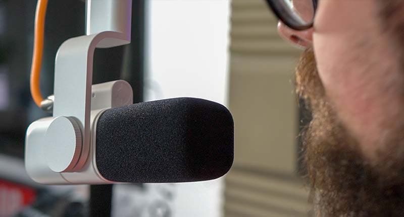 A close up shot of a man wearing glasses, speaking into a suspended Logitech Blue Sona mic.