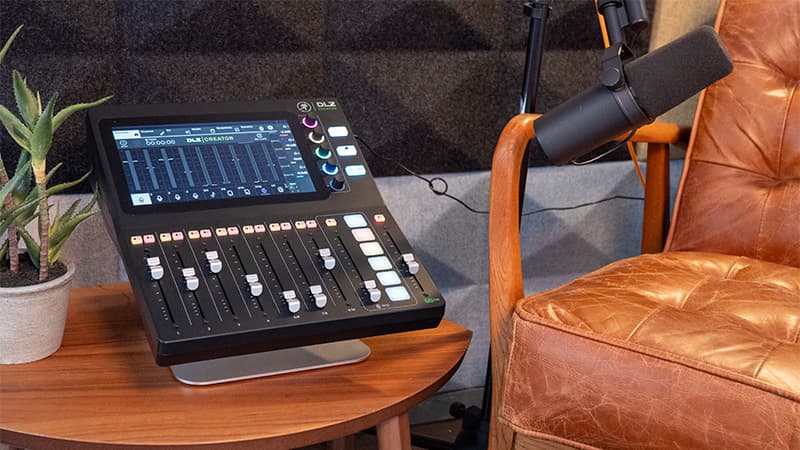 A Mackie DLZ Creator mixer on a stand, on a wooden coffee table. The mixer has a Shure SM7B microphone plugged in to it, and positioned over an empty leather chair.