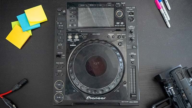 A Pioneer 2000-CDJ on a table, along with a video camcorder, post-its, pens and an XLR cable.