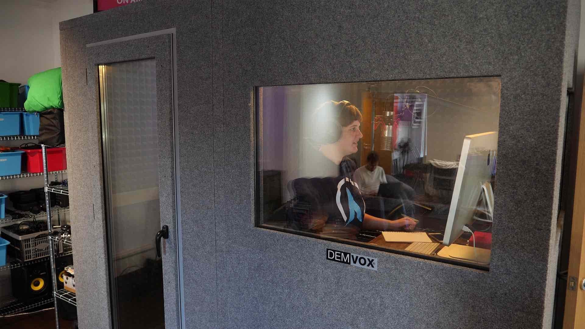 Image shows a stand alone radio booth, with one door and one window and a presenter in the booth speaking in a microphone.