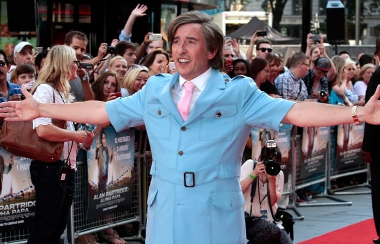 Image shows the character, Alan Partridge, wearing a blue short sleeve suit jacket and holding his arms out. Patridge, a white man in his 40s or 50s, is on the red carpet, and a small crowd is behind him looking in the other direction.