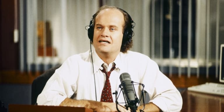Image shows a still of the character Frasier, from the show of the same name. Frasier is a white man in his 30s or 40s. He's wearing a white shirt and red tie, is sitting at a desk and talking in to a microphone whilst wearing headphones.