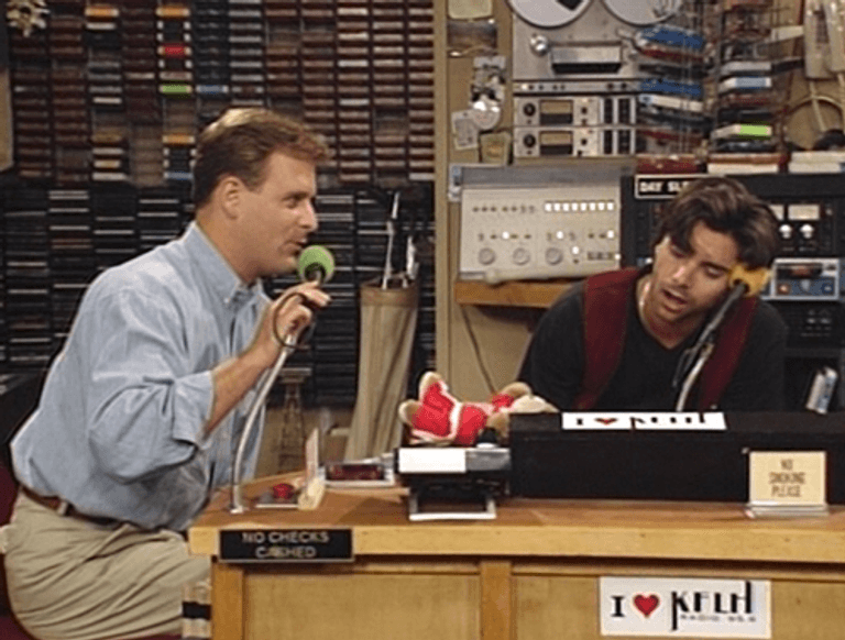 Image shows a scene from the TV show, Full House. The scene shows two white men in a radio studio, sat at a desk. One man is leaning over the desk excitedly and is talking into a microphone. While the other man is resting his head on his album and seemingly asleep.