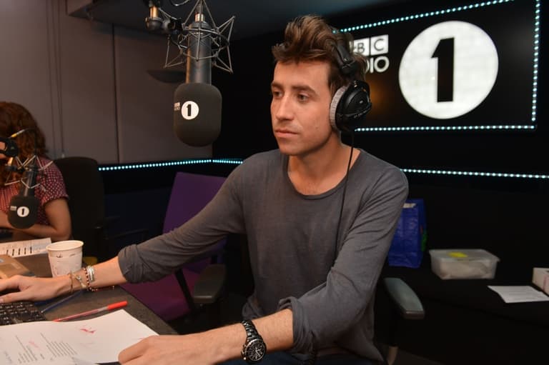 Radio 1 Presenter Nick Grimshaw is a white man in his 30s. He sits in the studio listening to a caller while recording a show, and wears headphones as he sits in front of the microphone in the studio.