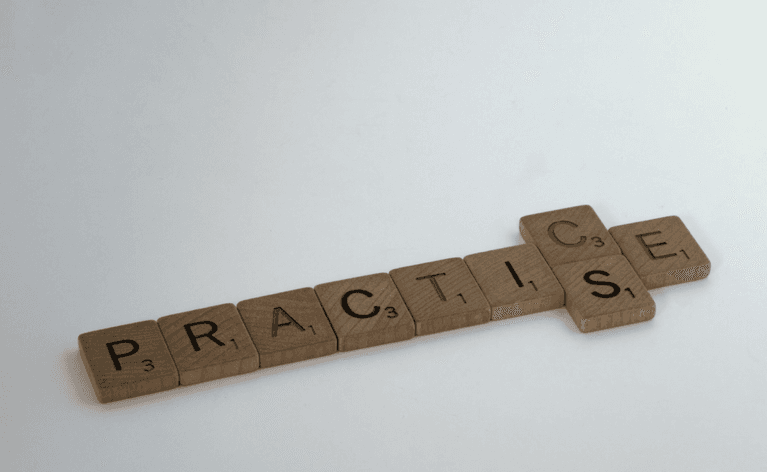 A wooden set of Scrabble tiles spells out 'Practice' with both the Americanised and Anglicised spellings.