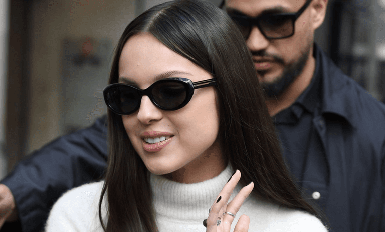 Pop singer Olivia Rodrigo smiles in a paparazzi photo. She is of Filipino descent and has long black hair and wears sunglasses. a