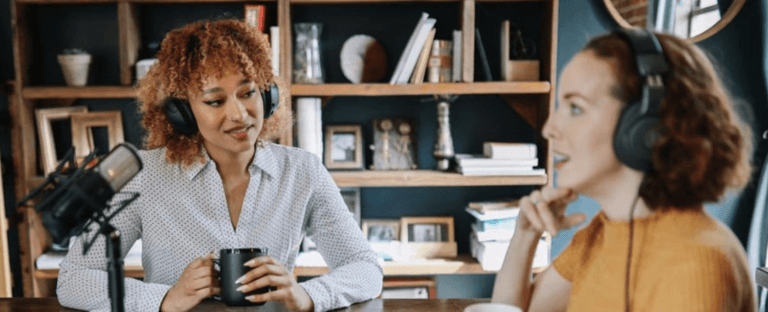 Two women sit opposite each other, one black and one white, and conduct a radio interview. Both have headphones on and one is leaning into the microphone while the other listens.