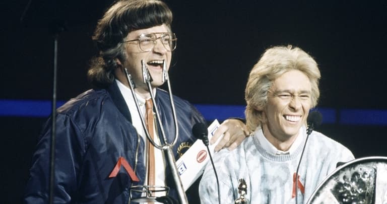 Image shows a still from the TV show, Smashie & Nicey. Smashie & Nicey are two white men laughing on stage. They are lit by a spotlight against a dark background.