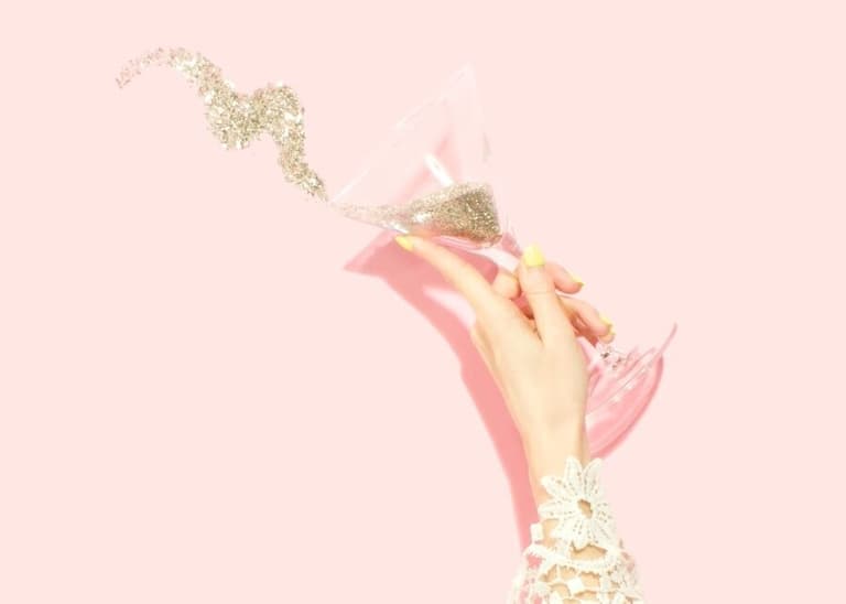 The background is pink. A celebrating person holds a martini glass and pours gold glitter from it.