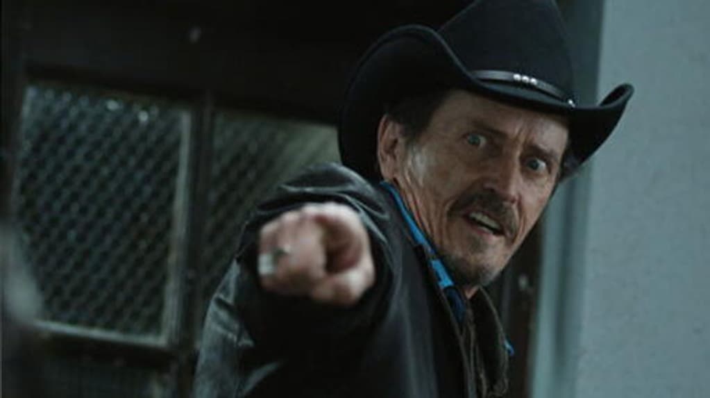 A white man with brunette hair, a 70s moustache, brown eyes and a black cowboy hat points at the camera. He has an expression of fear on his face.