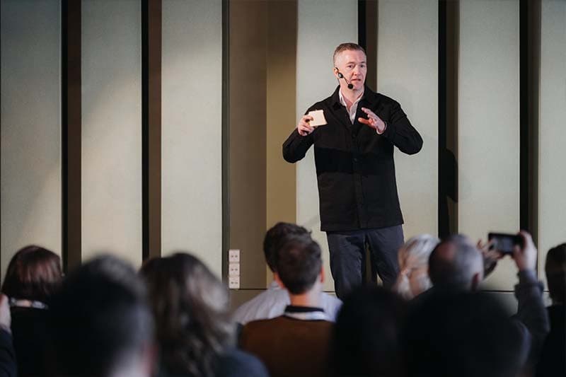 A white middle aged man delivering a talk on stage, with several rows of audience in front of him.