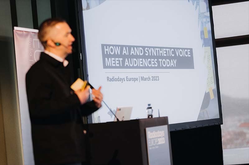 Image shows a white, middle-aged man - out of focus- talking on a stage, and in focus behind him is a screen that reads