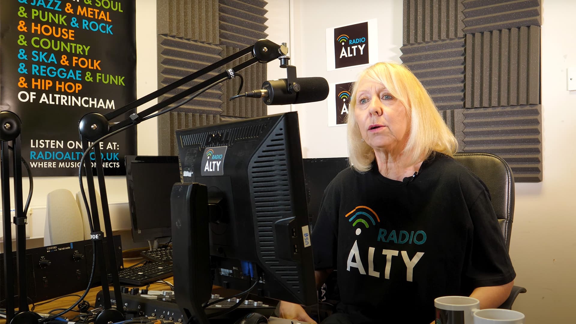 Annie, presenter of the Hot Buttered Soul show on Radio Alty, sits in front of a microphone as she speaks. She is a white, blonde older woman wearing a black and white Radio Alty t-shirt.