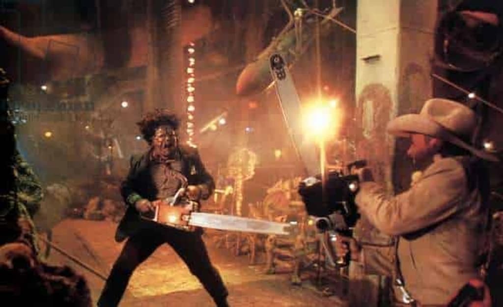 A movie set. The lighting is orangey yellow. On the right is a cameraman wearing a cowboy hat. In the middle is Leatherface, a scary figure in a mask, wielding a chainsaw at the camera.