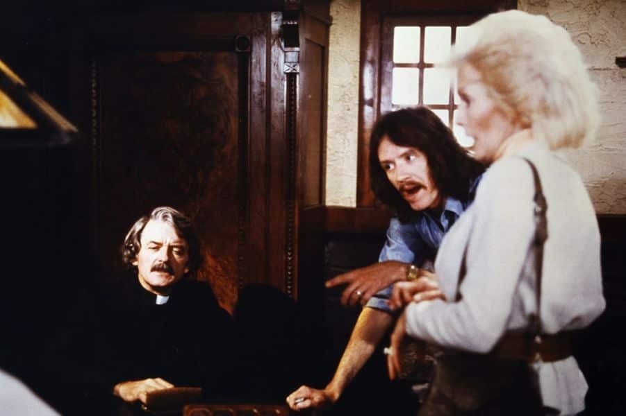 Three people stand in the lobby of a hotel. On the left is a white man with a moustache and gray hair, he is a priest. In the middle is a white man with long brown hair and a brown moustache. On the right is a white older woman with blonde hair.