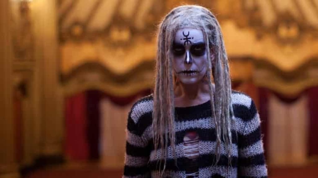 A white woman with blonde dreadlocks looks into the camera. She has white face makeup on with black eyes, like a skull, and has a satanic symbol on her forehead. She wears a striped black and white sweater. She is about 30.