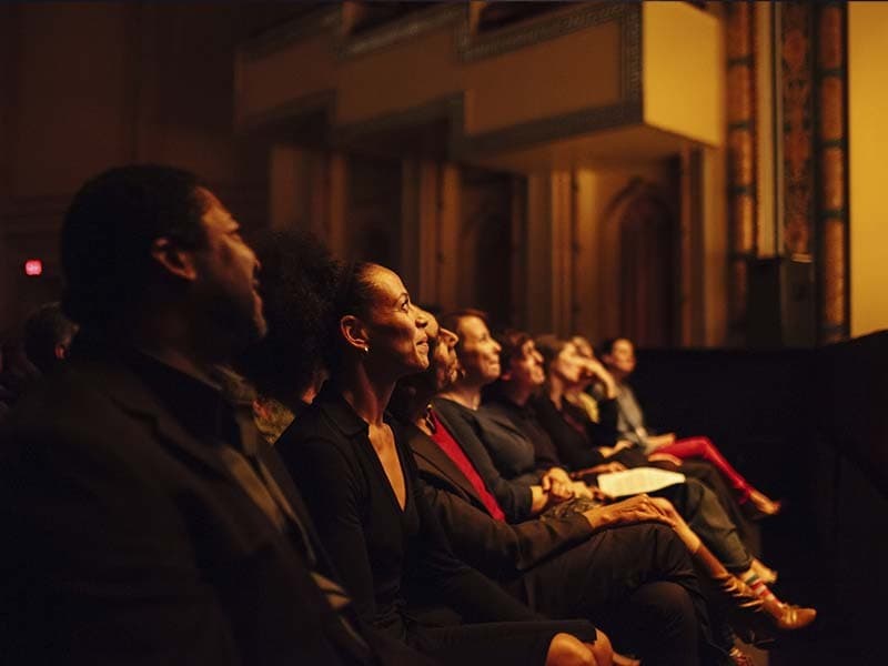 Image shows a row of an audience in a theatre, looking up at a stage that is out of shot.