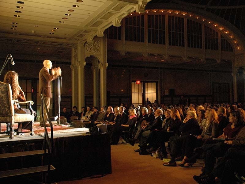 Image shows a man standing on stage, speaking in to a microphone and to a large audience.