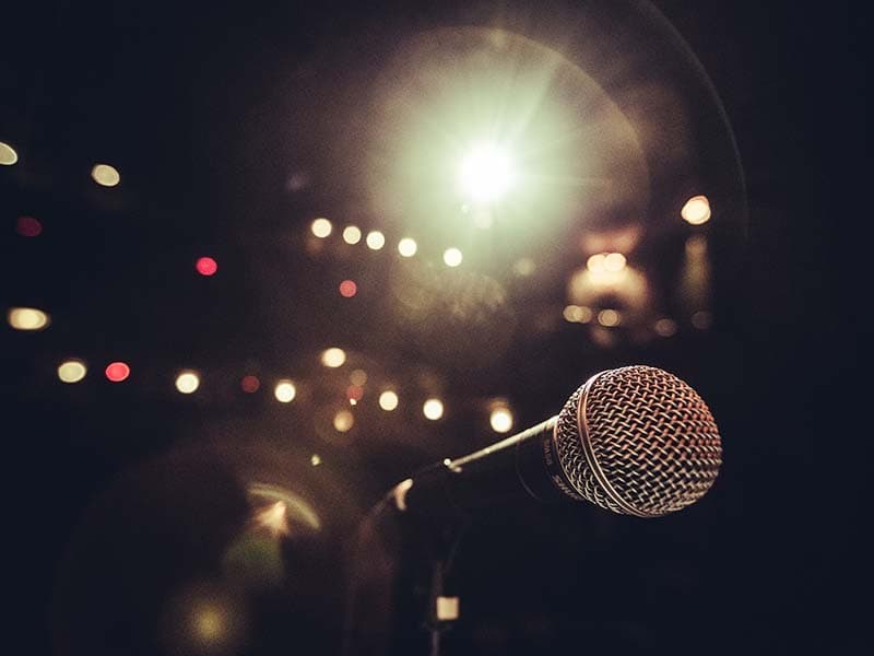 Image shows a close up of a microphone on a stand, in a theatre with the stage lights on.