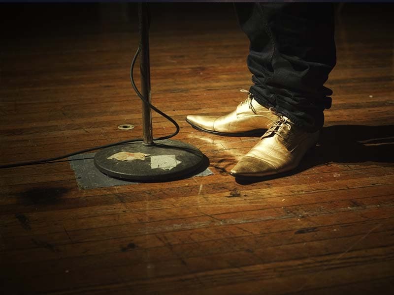 Image shows the feet of a person standing on stage next to a microphone stand. The person is wearing gold, pointy shoes and dark, loose trousers.