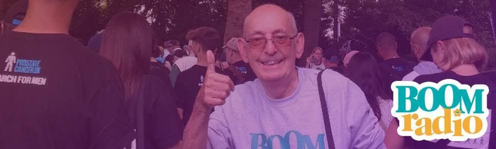 An older white man, stands with his back to a crowd, looks towards the camera and has his thumbs up. The Boom Radio logo is in the bottom-right hand corner.