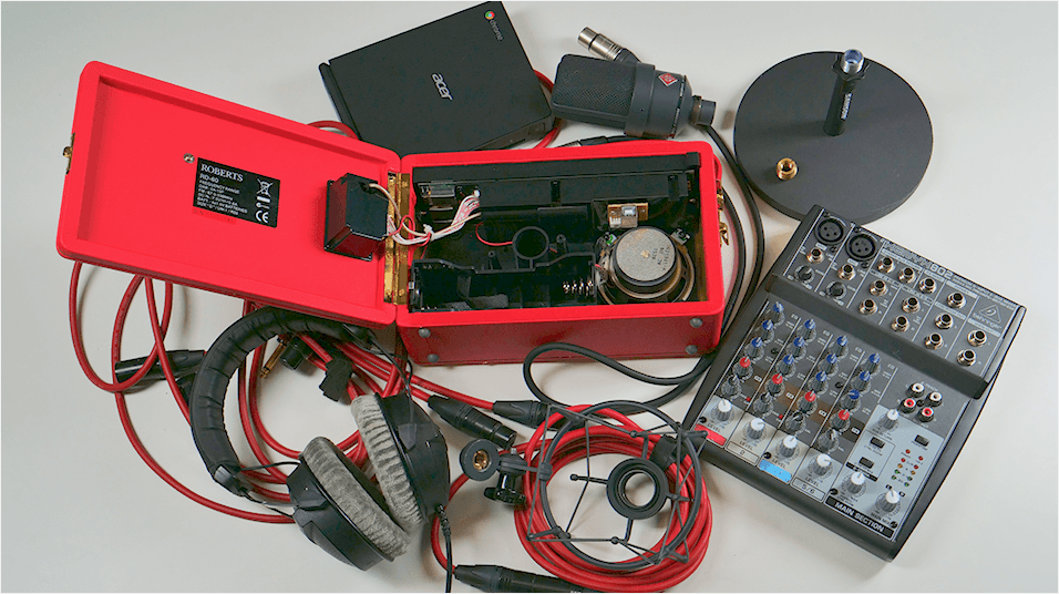 A birds-eye photo of a bundle of radio equipment, messily placed on a table. Equipment includes: an open radio set, a mixer, headphones, a shock mount, a microphone and cables.