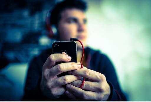 A man in his 20s, wearing headphones and listening to his iPhone. His iPhone and hands are in focus, and the rest of him is out of focus.