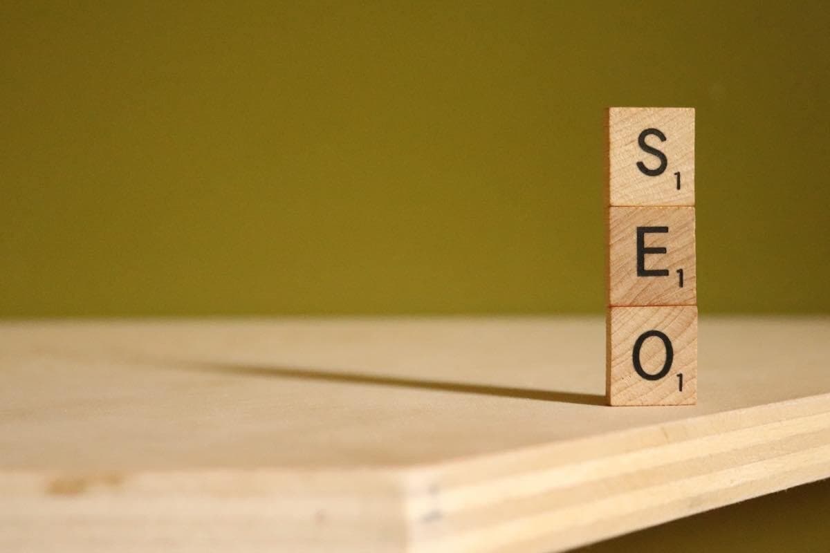 On a wooden table, three wooden scrabble tiles are stood up vertically on top of one another spelling out S E O.