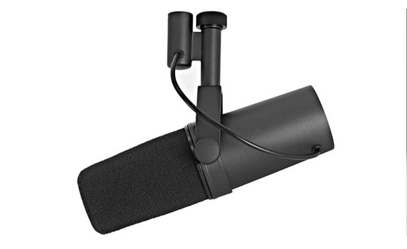 A Shure SM7B microphone hanging from a boom arm. The microphone is black and a capsule design and is against a white background.