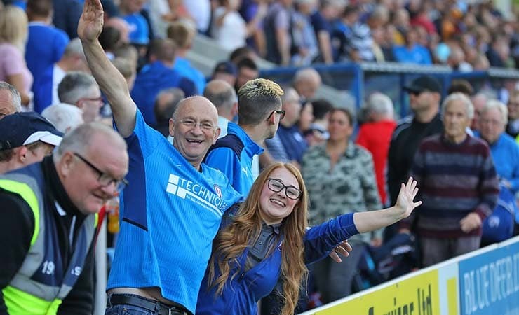 A crowd shot of the Chesterfield FC fans, with a man and a woman smiling, waving their arms and looking towards the camera.