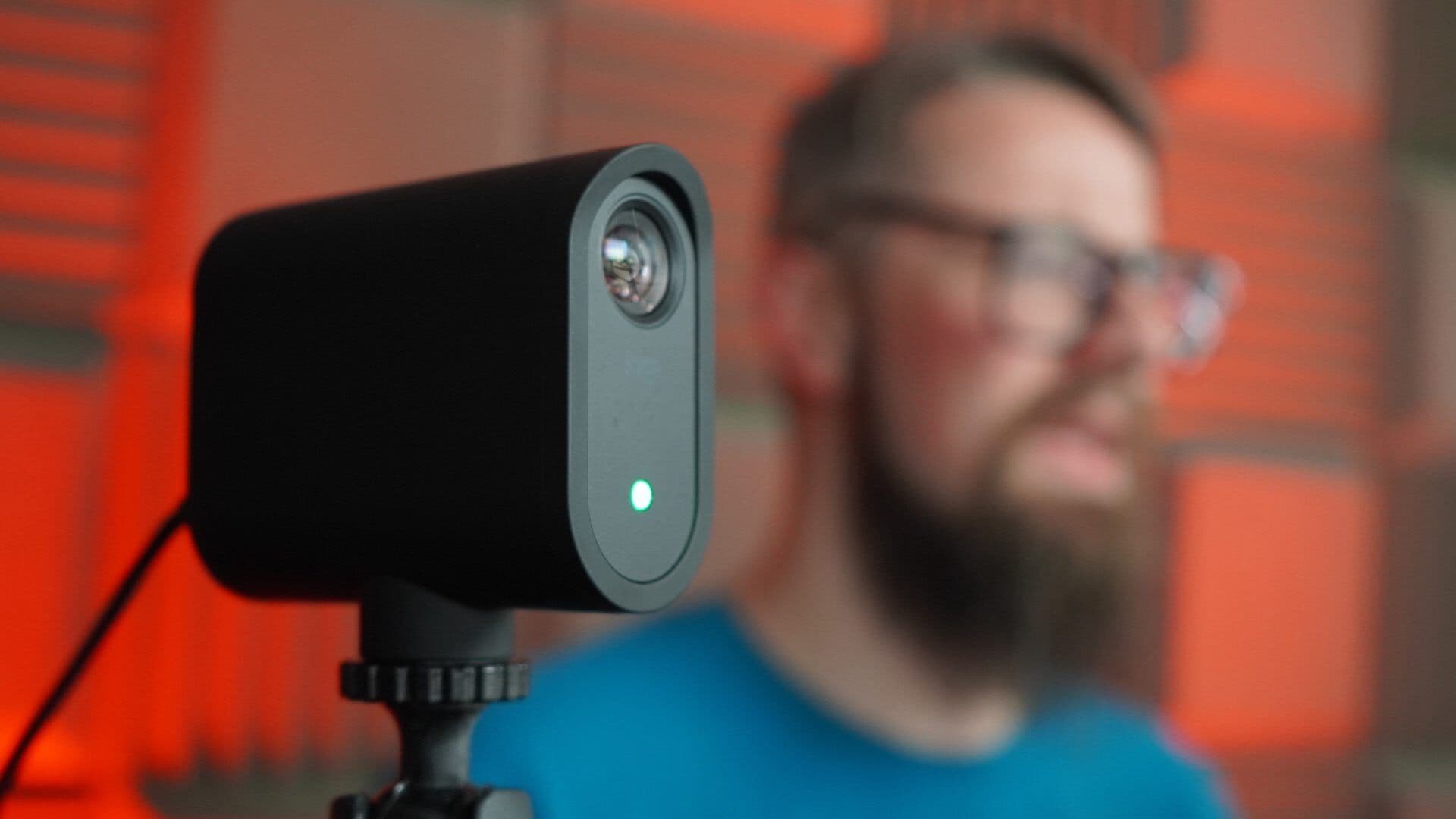 A Mevo camera in the foreground, pointing to the side and off-screen, with a man out of focus in the background.