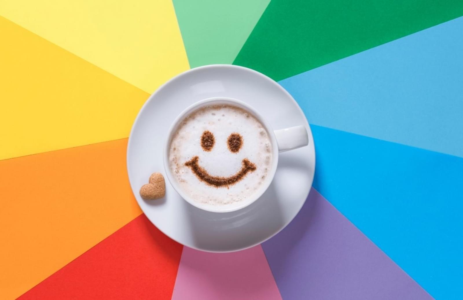 A cup of coffee with a smiley face dusted on top sits on a rainbow background