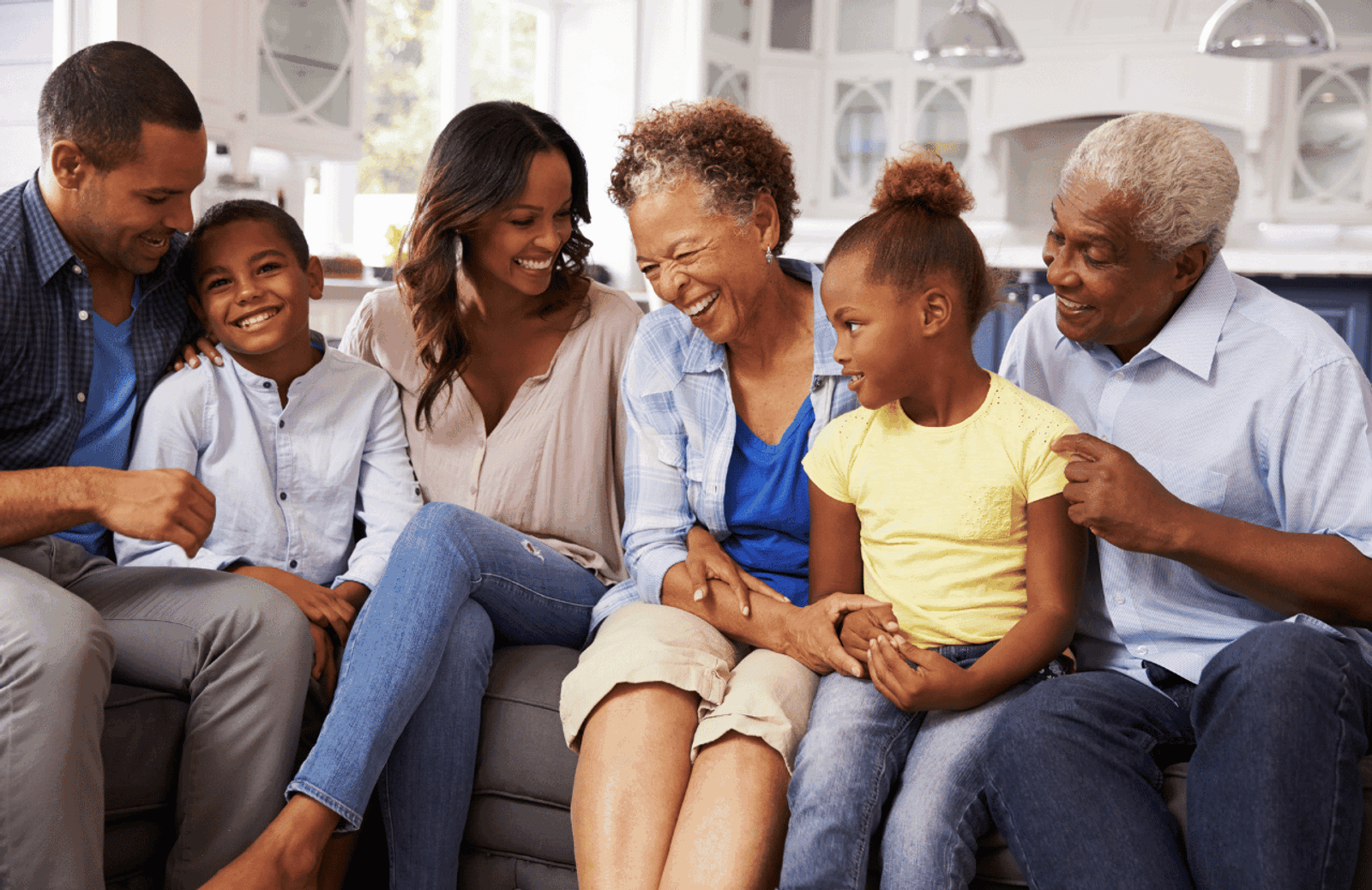 A Black multi-generational family sits on a couch together all smiling and laughing. There are two children, and four sets of adults, presumably the children's parents and grandparents.