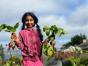 Sandhiya's daughter hols up two bunches of beets in each hand, a smile on her face and pigtails in her hair