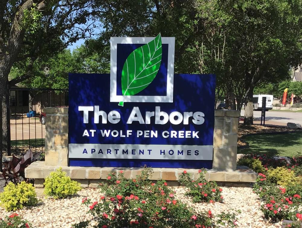 Entrance to Arbors at Wolf Pen Creek, featuring landscaped greenery and a blue and green sign under a blue sky.