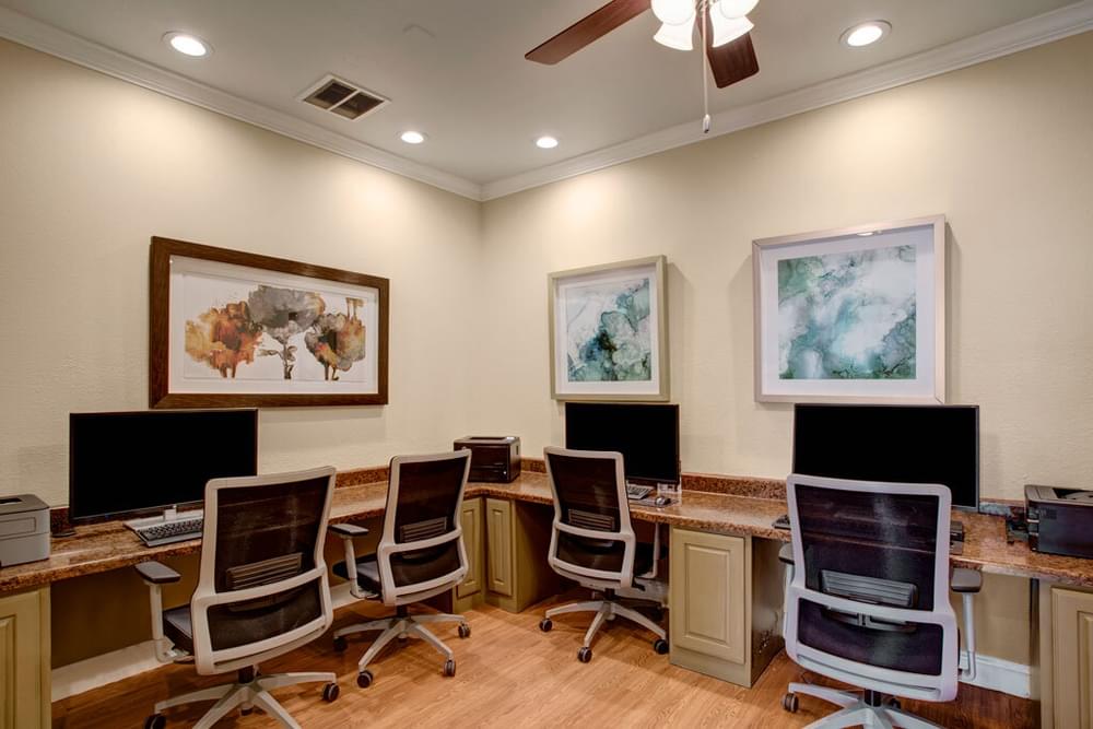 The Arbors at Wolf Pen Creek apartments Computer lab with four workstations and abstract art on beige walls. Ceiling fan above and wooden flooring.