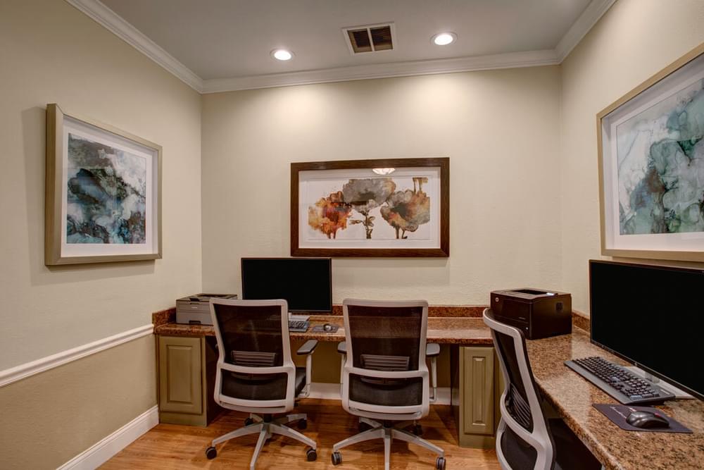 The Arbors at Wolf Pen Creek apartments Computer lab with four workstations and abstract art on beige walls. Ceiling fan above and wooden flooring.